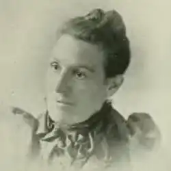 A white woman with dark hair dressed away from her face; she is wearing a dark dress with a high collar and puffed sleeves, with pins or ribbons on the chest