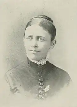 B&W portrait photograph of a woman with her hair in an up-do wearing a high-collared dark top with white fringe at the neckline.