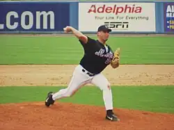 A man in a black baseball jersey and cap and gray baseball pants throws a baseball with his right hand.