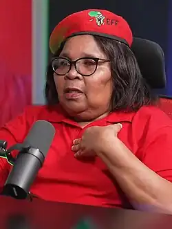 A woman seated at a table and behind a microphone, wearing a red beret and red polo shirt