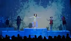 A color photograph of Marina performing live with backup singers while flowing their arms.