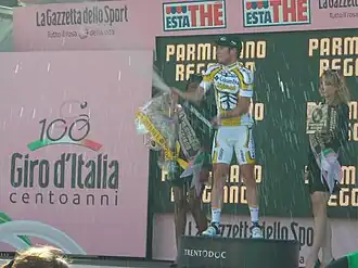A cyclist popping a champagne bottle on a podium, flanked by two women holding flowers and wine.