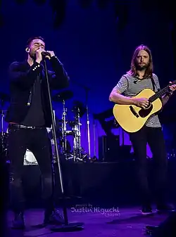 Two men performing on stage. One is wearing a black jacket with a black shirt and black denim jeans while singing into a microphone and the other one is wearing a black and white striped t-shirt and black jeans while playing the guitar.