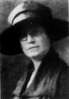 A white woman wearing a brimmed hat with a high crown, and glasses