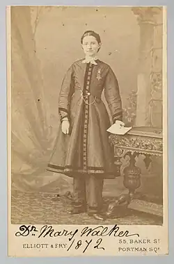 In this monochrome brown and white image, a white woman poses at center. Her complexion is white, with her hair braided and pinned close to her head. She wears a buttoned overcoat that ends at mid-calf with detailed banding on the hem and at wrists, and matching long pants beneath. She stands at center, on a brocade carpet, in front of a painted backdrop, her left hand rests on a carved wooden table, holding a document, her right hand rests in front of her coat. The printed image is mounted on a white card, which is annotated at the bottom "Dr. Mary Walker 1872" in manuscript ink, above and between the printed name and address of the photograph studio [55 Baker Street, Portman Square]