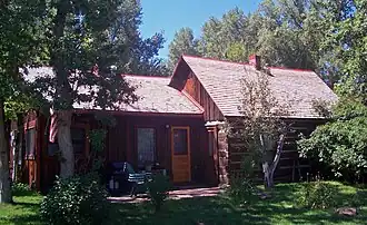 An unpainted dark brown wooden house amid tall trees in two sections. The taller one, on the right, has a pointed roof with a small brick chimney rising from the middle.