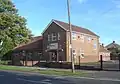 Three-quarter view of a vernacular-style brick building in two parts, behind a black fence and flanked by a grass area and a car park. The near part has two storeys with three windows above the entrance door and irregularly placed windows on the side wall. The far section has a long, steep roof and one storey.