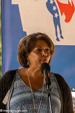 A woman stands at a podium with a microphone in front of her. She is wearing a blue blouse and a dark shawl.