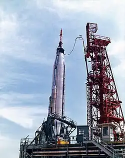 A smooth cylindrical rocket, with a small conical spacecraft at its tip, with its base held by a metal cradle. It is connected by a long cable to a tall framework structure to its right.