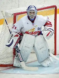 An ice hockey player standing partially crouched in goals. He is wearing a helmet, gloves and leg pads and a white uniform.