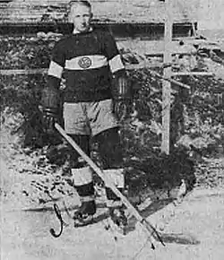 A man stands outside in skates, wearing a sweater with a team logo on it, and wears gloves and holds an ice hockey stick.