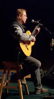 A fair-haired man sitting on a stool playing a guitar