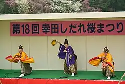 Three men in traditional Japanese clothes, one with fan, two with drums on a stage.