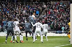 Several soccer players face away from the camera against a background with a full crowd; one player has jumped towards the in-flight ball while the goalkeeper looks on behind a goalpost.