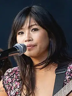 Beabadoobee behind the mic, playing guitar, made-up and wearing a floral blouse.