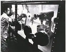 A black and white photograph of a man wearing a full body HAZMAT suit, standing on the left end, operating some machinery fully covered behind glass