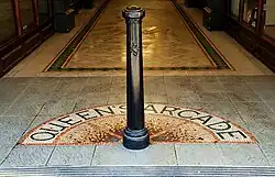 Close-up photograph of a black Victorian-style bollard in the centre of the arcade walkway. A semi-circular mosaic on the floor spells out the letters "Queen's Arcade" around the bollard.