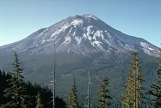 Large volcanic mountain deformed by large cracks and bulges on its slopes, seen from a ridge across a thickly forested valley. A summit crater is partially seen.