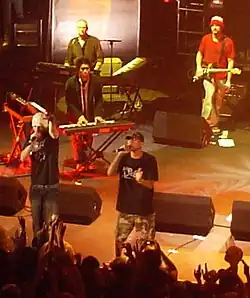 All-male group on stage, containing two MCs, two keyboardists, and a guitarist. Some of the crowd with hands in the air present in the foreground