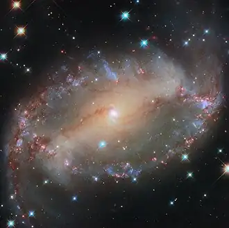 An oval-shaped spiral galaxy. Its core is a compact, glowing blue spot. A bright bar of light, lined with dark reddish dust, extends horizontally to the edge of the disc. A spiral arm emerges from each end of the bar and follows the edge of the disc, lined with blue and red glowing patches of stars, to the opposite end and a little off the galaxy. Blue stars are scattered between us and the galaxy.