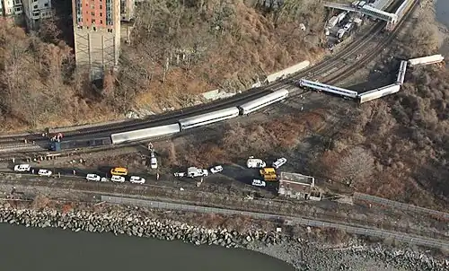 An aerial view of a wooded area between two watery areas in late autumn, with two railroad tracks forking to the top near the left and one following the water's edge. Several silvery train cars and a locomotive are lying on their sides off the tracks to the top; there are many parked yellow and white trucks and other vehicles between the two tracks.