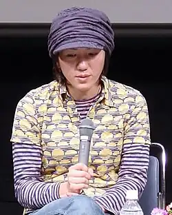 A Japanese woman wearing a newsboy cap and patterned shirt, holding a microphone.