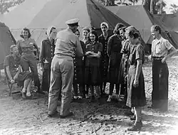A photograph from 1945, taken outdoors, in front of a series of large tents; about 12 young white women, mostly in denim dresses, are standing and sitting; a man in uniform has his back to the camera.