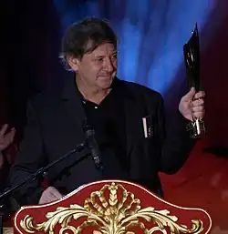 middle-aged white male wearing a dark suit, standing at a podium, holding trophy, looking left of camera