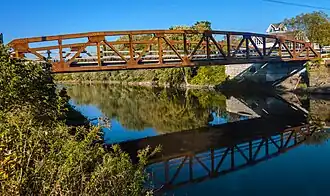 A brown metal bridge with low arches on each side, crossing brown water that reflects it, seen from one side under a blue sky. Across the water is a house, with trees lining the water's edge beyond the bridge.