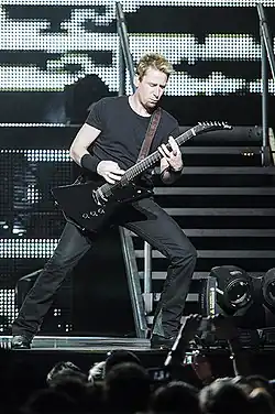 A blonde man wearing a dark T-shirt and dark jeans playing a guitar on a stage.