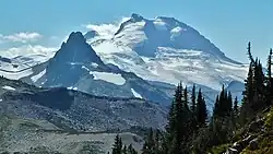 A prominent, glaciated mountain overlooking a rocky peak on the left and a forested slope on the right.
