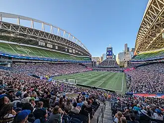 The lower grandstand of a stadium filled with people during a game, while the upper stands are covered by decorative tarps. The stadium has distinctive trusses for support.