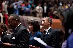 Obama and his wife standing in a crowded Church, looking forward, with their mouths open mid-sentence while reciting a prayer
