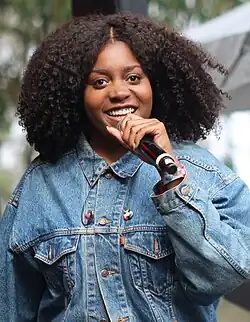 Noname onstage smiling, holding a microphone