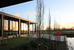 A photo showing the outside of the David Kato hub overlooking the pond. The photo was taken at sunset on a January afternoon, the sun is reflecting in the glass of the hub with contrasting black metal pillars.