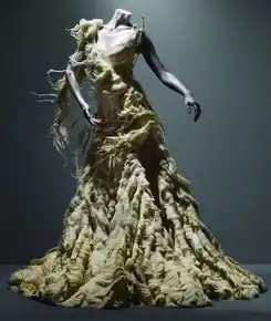 Full-length beige sleeveless dress displayed on a mannequin. The full skirt is made from numerous layers of thin fabric stacked to appear like an oyster shell. The dress has been intentionally distressed and shredded.