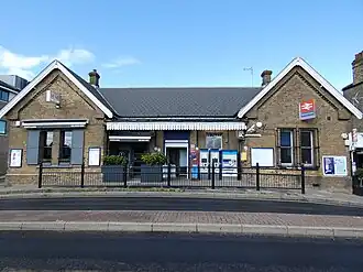 A fairly large brick building with a tiled roof and chimneys as viewed from across the street. In the centre of the building there is a doorway with a sign above that says "Welcome to Palmers Green", as well as a shop on the left and ticket machines on the right, all of which are underneath a canopy structure.