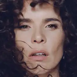 A close-up portrait of a woman with curly dark brown hair, an intense yet serene expression, slightly parted lips, deep brown eyes, well-defined eyebrows, a natural complexion with a subtle glow, and strands of hair framing her face against a blurred background with soft, even lighting.