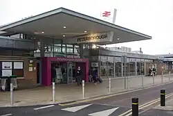 A modern glass building with a pink doorway, a sign on the top of which says "PETERBOROUGH STATION". On each side the glass becomes brick wall after a few windows. A very large flat metal canopy sits above the entranceway; a parallelogram intersects this canopy perpendicular to the entranceway, with "PETERBOROUGH" below the canopy and a double arrow above.