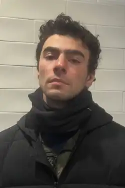 Headshot of Luigi Mangione in a McDonalds. He is a young, white man with short, curly, black hair, large eyebrows, a defined jaw, and light stubble beard. He is wearing a large black jacket and a black scarf. He is looking at the viewer with a calm but unsmiling expression.