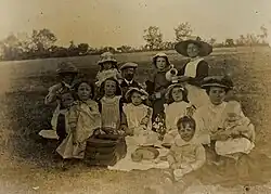 monochrome photograph of early 20th century group of thirteen adults and children seated with a picnic on a grass slope with hedge and trees in the background