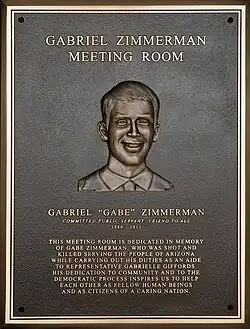 A bronze plaque. Engraved in the center of the place is the head and shoulders of Gabriel Zimmerman. The plaque reads: "GABRIEL ZIMMERMAN MEETING ROOM GABRIEL “GABE” ZIMMERMAN COMMITTED PUBLIC SERVANT; FRIEND TO ALL 1980-2011 THIS MEETING ROOM IS DEDICATED IN MEMORY OF GABE ZIMMERMAN, WHO WAS SHOT AND KILLED SERVING THE PEOPLE OF ARIZONA WHILE CARRYING OUT HIS DUTIES AS AN AIDE TO REPRESENTATIVE GABRIELLE GIFFORDS. HIS DEDICATION TO COMMUNITY AND TO THE DEMOCRATIC PROCESS INSPIRES US TO HELP EACH OTHER AS FELLOW HUMAN BEINGS AND AS CITIZENS OF A CARING NATION".