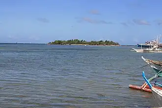 Small low green island in blue sea, at about 600 m, with several palm trees on top. To right prows of four bangkas – local boats for fishing or diving.