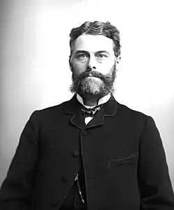 B & W medium-shot photographic portrait of Edward Drinker Cope, paleontologist, c. 1880, in early middle age with dark hair and greying, scruffy full beard; wearing a dark coat and soft bow tie.