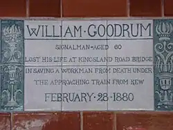 A tablet formed of six standard sized tiles, bordered by green flowers in the style of the Arts and Crafts movement. The tablet reads "William Goodrum signalman aged 60 lost his life at Kingsland road bridge in saving a workman from death under the approaching train from Kew February 28, 1880".
