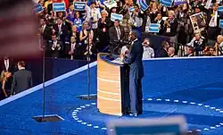 A man, wearing a suite, standing behind two transparent, glass teleprompters