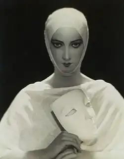 Black and white image of a woman wearing white, from the chest up. Her hair is covered with a white wrap and she holds a white mask.