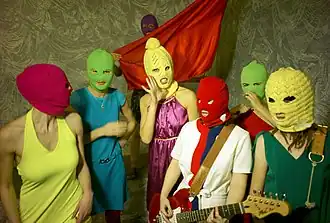 7 women with bright colored clothes and multicolored knit ski masks over their faces. A woman at the center holds a guitar and one at the back holds a piece of red fabric.