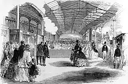 An artist's impression of the inside of a train station crowded with people underneath a long roof. One group of these is obviously identifiable as Queen Victoria , Prince Albert , and one of their chlidren. Two men bow their heads opposite them.