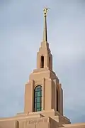 The spire of the Red Cliffs Utah Temple dominates the view of a portrait shot. A multilevel spire has rounded windows on the sides, along with a golden statue at the top of the angel Moroni, against a backdrop of a blue sky.